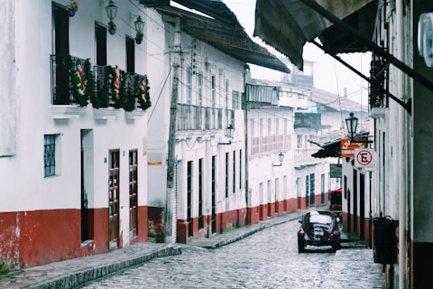 A cobblestone street in a quaint town is lined with traditional white buildings, featuring red accents on their lower halves. Some buildings have balconies adorned with garlands. A vintage car is parked along the road near a sign indicating parking restrictions. The area appears calm and quiet, with hanging lanterns adding a rustic touch.