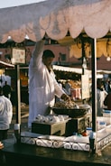 A vendor wearing a white coat stands at a market stall selling nuts. The stall has an ornate metal railing and is laden with bowls and containers. In the background, blurred figures move through a busy marketplace.