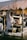 A vendor wearing a white coat stands at a market stall selling nuts. The stall has an ornate metal railing and is laden with bowls and containers. In the background, blurred figures move through a busy marketplace.