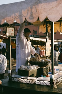 A vendor wearing a white coat stands at a market stall selling nuts. The stall has an ornate metal railing and is laden with bowls and containers. In the background, blurred figures move through a busy marketplace.