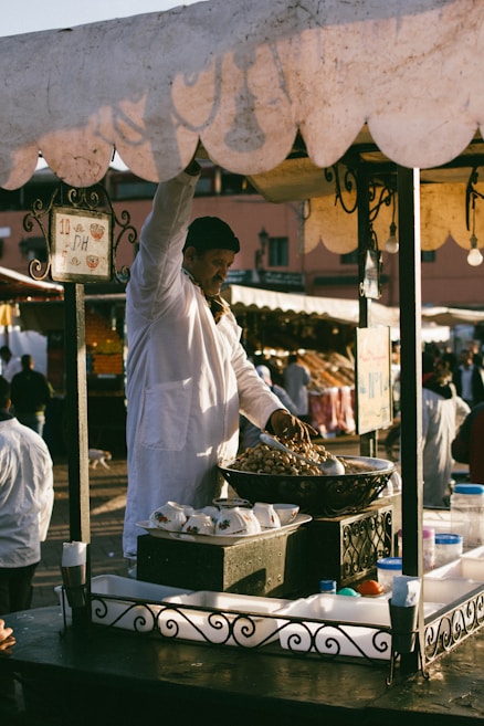 A vendor wearing a white coat stands at a market stall selling nuts. The stall has an ornate metal railing and is laden with bowls and containers. In the background, blurred figures move through a busy marketplace.