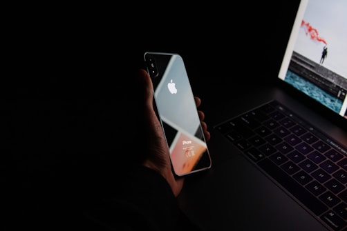 A hand holding a reflective iPhone positioned over a laptop keyboard, with the screen displaying an image. The dim lighting creates a moody atmosphere with a focus on the sleek design of the devices.