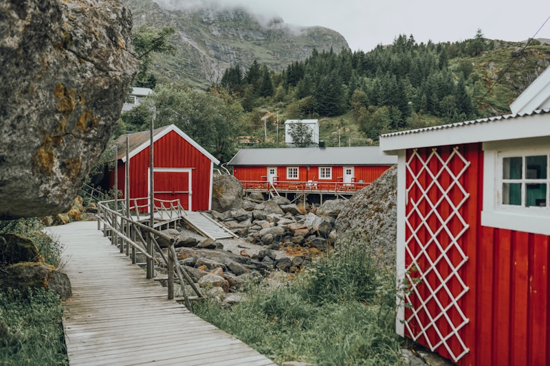 A picturesque scene featuring traditional red wooden cabins with white trim, situated in a rocky landscape. A wooden boardwalk leads past the cabins, surrounded by lush greenery and rugged mountains in the background. The sky appears overcast, adding to the serene atmosphere.