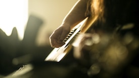 A close-up view of a person strumming the strings of an acoustic guitar, with soft ambient lighting creating a warm and intimate atmosphere. The focus is on the hand and the guitar strings, while the background is blurred, enhancing the sense of depth.
