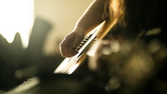 A close-up view of a person strumming the strings of an acoustic guitar, with soft ambient lighting creating a warm and intimate atmosphere. The focus is on the hand and the guitar strings, while the background is blurred, enhancing the sense of depth.