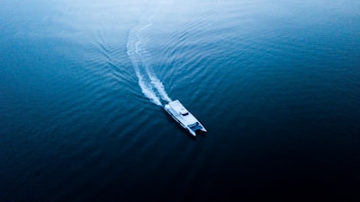 A lone catamaran glides smoothly across a vast expanse of calm blue water, leaving a trail of gentle ripples behind. The scene is serene and tranquil, capturing the peacefulness of open waters.