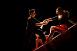 A dramatic and dynamic scene featuring dancers in motion on a dimly lit stage. A man extends his hand towards a woman who is poised gracefully on a sloped surface, wearing a flowing red skirt. Their expressions are intense and focused, capturing a moment of connection.