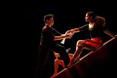 A dramatic and dynamic scene featuring dancers in motion on a dimly lit stage. A man extends his hand towards a woman who is poised gracefully on a sloped surface, wearing a flowing red skirt. Their expressions are intense and focused, capturing a moment of connection.