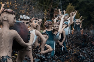 A line of whimsical ceramic or clay figures with raised arms are set in a garden of dark foliage. The figures have detailed facial features and varying expressions, adorned with decorative elements like colored beads or stones in their hair. The setting is outdoors, with surrounding plants and trees creating a natural backdrop.
