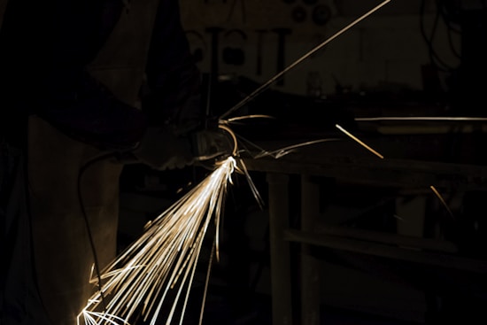Sparks fly as an angle grinder is used in a dimly lit workshop. The person operating the tool is partially visible, emphasizing the action and the bright, glowing sparks against a dark background.