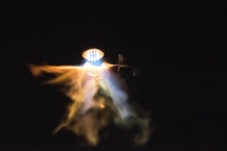 A bright, glowing light source illuminates a blend of swirling smoke or mist, presenting a dynamic mixture of yellow, orange, and blue hues against a dark background.