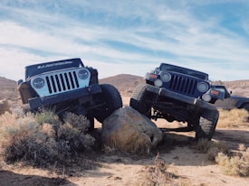 Two off-road vehicles are positioned on a rocky terrain, each with one wheel elevated on a boulder, showcasing their suspension capabilities. The background features a desert landscape under a clear blue sky.