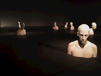 Several human mannequins are partially submerged in a dark, reflective surface. The scene has a surreal, almost haunting quality with dim lighting and shadows. The mannequins are positioned in rows, creating an impression of uniformity and contemplation.