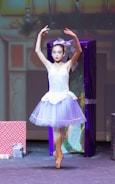 A ballerina in a white and light purple tutu performs on stage en pointe, surrounded by large, colorful wrapped presents. The background is softly lit, suggesting a festive setting.