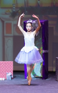 A ballerina in a white and light purple tutu performs on stage en pointe, surrounded by large, colorful wrapped presents. The background is softly lit, suggesting a festive setting.