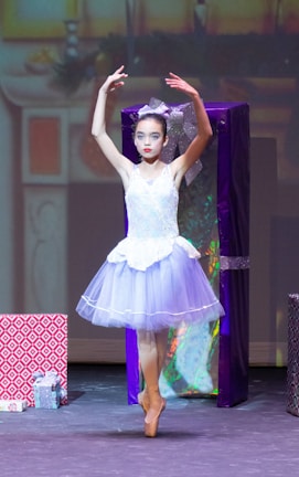 A ballerina in a white and light purple tutu performs on stage en pointe, surrounded by large, colorful wrapped presents. The background is softly lit, suggesting a festive setting.