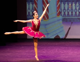 A ballerina performs an elegant pose on stage, wearing a vibrant pink tutu adorned with gold embellishments. The background features a colorful, theatrical set design with a balcony and striped columns, while the stage is lit with purple hues.