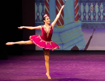 A ballerina performs an elegant pose on stage, wearing a vibrant pink tutu adorned with gold embellishments. The background features a colorful, theatrical set design with a balcony and striped columns, while the stage is lit with purple hues.