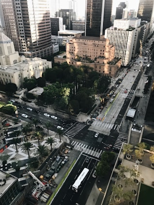 An aerial view of an urban area featuring several high-rise buildings, intersecting roads, and a well-maintained park with green trees. The streets are bustling with vehicles, including a visible white bus, and pedestrians can be seen crossing. The buildings exhibit varying architectural styles and heights, with sunlight casting shadows across the scene.