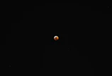 A small, reddish-orange celestial body is surrounded by a vast expanse of black space, likely depicting a lunar eclipse or a similar astronomical event.