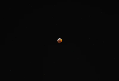 A small, reddish-orange celestial body is surrounded by a vast expanse of black space, likely depicting a lunar eclipse or a similar astronomical event.