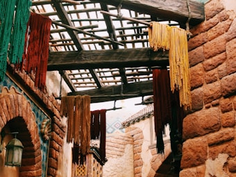 A rustic setting featuring bundles of colorful dyed yarn hanging from a wooden framework. The walls are constructed of reddish-brown stone, and a decorative lantern hangs to the side. The scene suggests a traditional or historical market atmosphere.