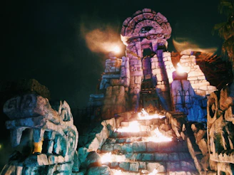 An ancient, Mayan-style stone structure with intricate carvings and a large, menacing face sculpture dominates the foreground. The scene is dramatically lit with torches and spotlights casting an eerie glow over the stone steps leading up to the central archway, and the sky is dark with a hint of smoke or mist.