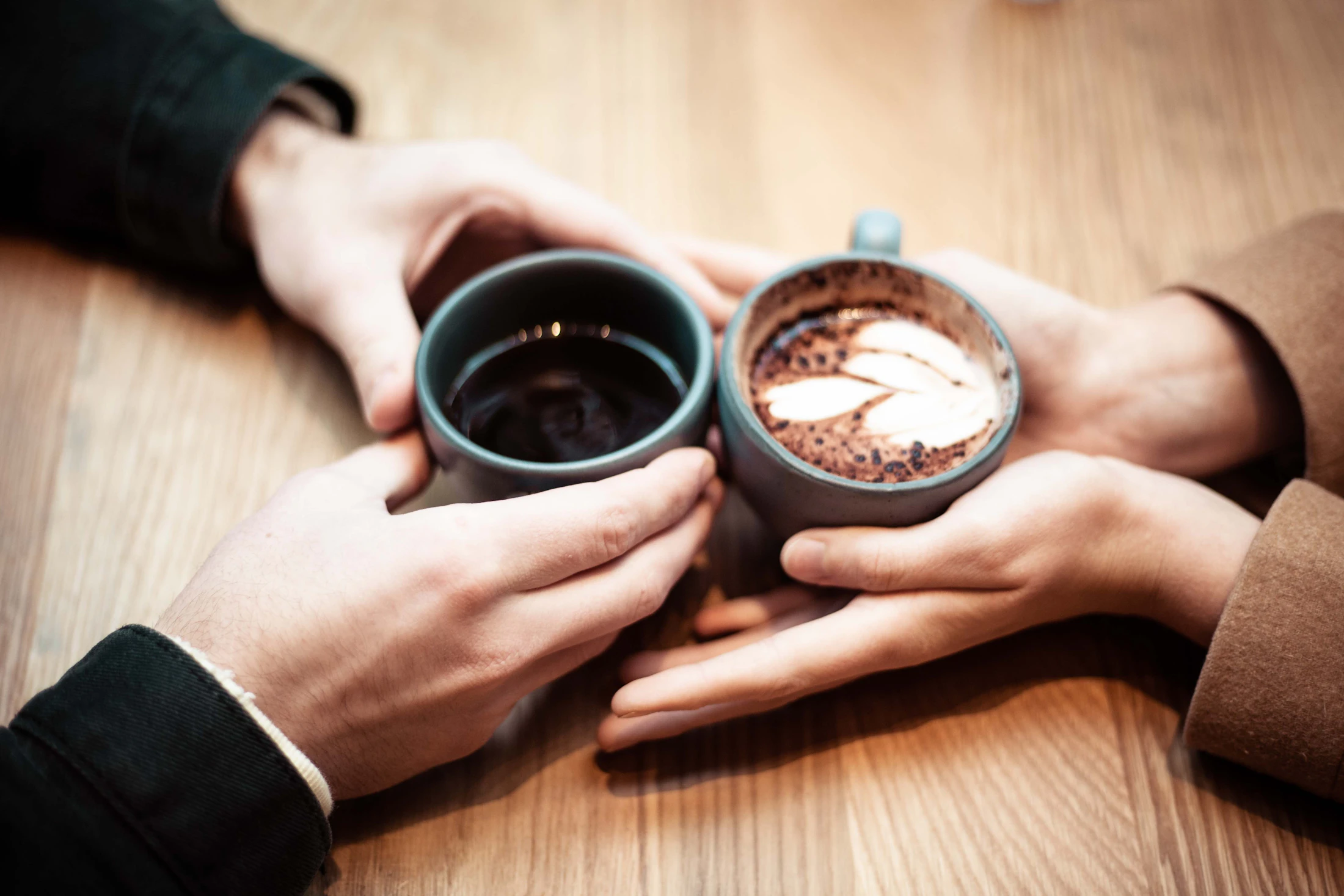 Two people passing warm mugs of coffee to each other across a wooden table