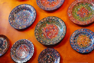 A collection of vibrantly painted ceramic plates displayed on an orange textured wall. Each plate features intricate floral patterns in a variety of colors, including blue, red, green, and orange, highlighting detailed craftsmanship.