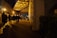 A dimly lit walkway beneath an urban bridge, with several people walking in both directions. Streetlights cast long shadows on the pavement, and the area is bordered by a brick wall and greenery. The bridge overhead features a grid-like metal structure.