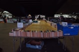 A makeshift outdoor bookstall displays a wide array of books organized in rows on tables. Signs indicate different genres like Sci-Fi and Fantasy. The setting is under a bridge with graffiti-covered walls and city lights in the background, creating a dimly lit yet vibrant urban atmosphere.