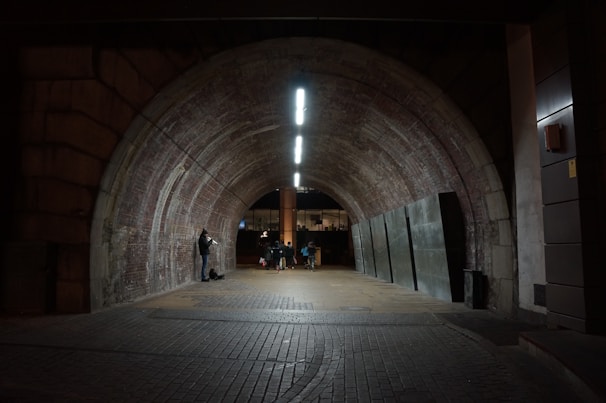 A dimly lit tunnel with a brick ceiling creates an atmospheric setting. A musician plays a brass instrument on the left side, while a group of people walk toward the far end. Modern lighting enhances the textured walls, and an industrial atmosphere prevails.