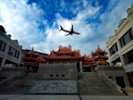 A traditional Asian temple with ornate, red-tiled rooftops stands prominently beneath a mostly cloudy sky. An airplane flies overhead, providing a juxtaposition of modern technology against historical architecture. The temple is flanked by other buildings with similar architectural styles.