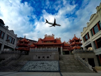 A traditional Asian temple with ornate, red-tiled rooftops stands prominently beneath a mostly cloudy sky. An airplane flies overhead, providing a juxtaposition of modern technology against historical architecture. The temple is flanked by other buildings with similar architectural styles.