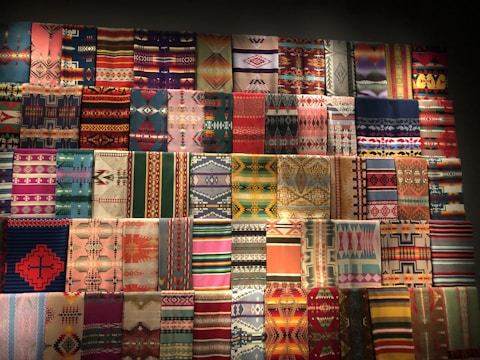 A collection of colorful, patterned textiles are arranged in a grid on a wall. The textiles feature a diverse range of geometric designs and vibrant hues, including stripes and intricate motifs.
