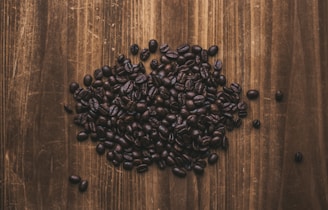 A pile of dark roasted coffee beans is spread out on a rustic wooden surface. The texture of the wooden surface has visible grains and patterns, contrasting with the shiny, uneven shapes of the coffee beans.