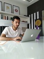 A man is focused on his laptop in a modern office setting with white walls and black-striped decor. Above him are three framed abstract art pieces, and to the side is a bookshelf filled with various books. A target board with darts is mounted on the wall, and a purple bottle is placed on the white glossy table.