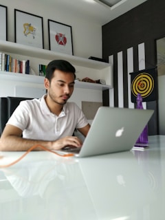 A man is focused on his laptop in a modern office setting with white walls and black-striped decor. Above him are three framed abstract art pieces, and to the side is a bookshelf filled with various books. A target board with darts is mounted on the wall, and a purple bottle is placed on the white glossy table.