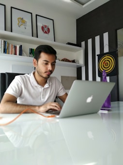 A man is focused on his laptop in a modern office setting with white walls and black-striped decor. Above him are three framed abstract art pieces, and to the side is a bookshelf filled with various books. A target board with darts is mounted on the wall, and a purple bottle is placed on the white glossy table.