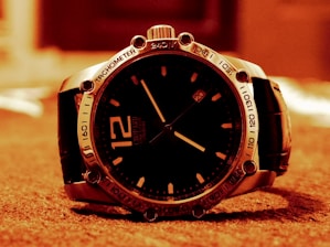 A close-up image of a luxury wristwatch resting on a soft surface. The watch has a round, dark dial with sleek hands and a prominent number 12. The bezel has engraved markers and numbers, and the strap appears to be leather. The lighting casts a warm, sepia-tone over the entire scene.