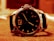 A close-up image of a luxury wristwatch resting on a soft surface. The watch has a round, dark dial with sleek hands and a prominent number 12. The bezel has engraved markers and numbers, and the strap appears to be leather. The lighting casts a warm, sepia-tone over the entire scene.