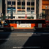A bright orange city bus with advertisements displayed on its side, driving along a city street in front of a commercial building with store signs. The scene includes a shadow cast on the road and trees lining the street.