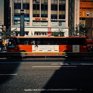 A bright orange city bus with advertisements displayed on its side, driving along a city street in front of a commercial building with store signs. The scene includes a shadow cast on the road and trees lining the street.