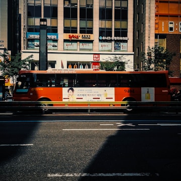 A bright orange city bus with advertisements displayed on its side, driving along a city street in front of a commercial building with store signs. The scene includes a shadow cast on the road and trees lining the street.