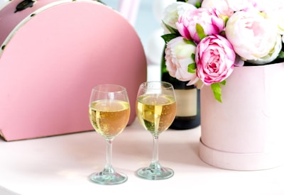 Two glasses of sparkling wine are positioned on a light-colored surface beside a bouquet of pink and white peonies in a pastel cylindrical container. A partial view of a pink, rounded box or suitcase is visible in the background, adding to the elegant and celebratory setting.