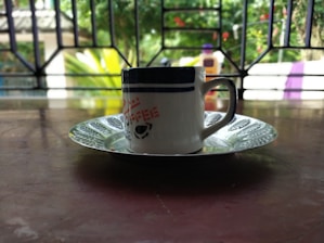 A ceramic mug with a patterned design sits on a decorative saucer, placed on a wooden table. In the background, there is a view of greenery through a window with wrought iron bars.