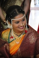 A woman dressed in traditional Indian attire with intricate jewelry, including a pearl headdress and a nose ring. The sari is a combination of rich maroon and vibrant orange with gold embroidery. Her hair is adorned with white flowers, and she displays a warm smile.
