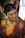 A woman dressed in traditional Indian attire with intricate jewelry, including a pearl headdress and a nose ring. The sari is a combination of rich maroon and vibrant orange with gold embroidery. Her hair is adorned with white flowers, and she displays a warm smile.