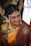 A woman dressed in traditional Indian attire with intricate jewelry, including a pearl headdress and a nose ring. The sari is a combination of rich maroon and vibrant orange with gold embroidery. Her hair is adorned with white flowers, and she displays a warm smile.