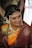 A woman dressed in traditional Indian attire with intricate jewelry, including a pearl headdress and a nose ring. The sari is a combination of rich maroon and vibrant orange with gold embroidery. Her hair is adorned with white flowers, and she displays a warm smile.